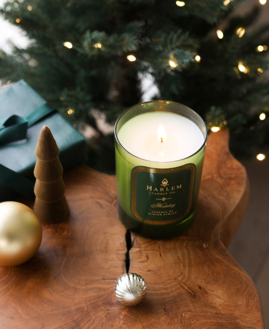 Green candle with a label on a wooden surface in front of a decorated Christmas tree.