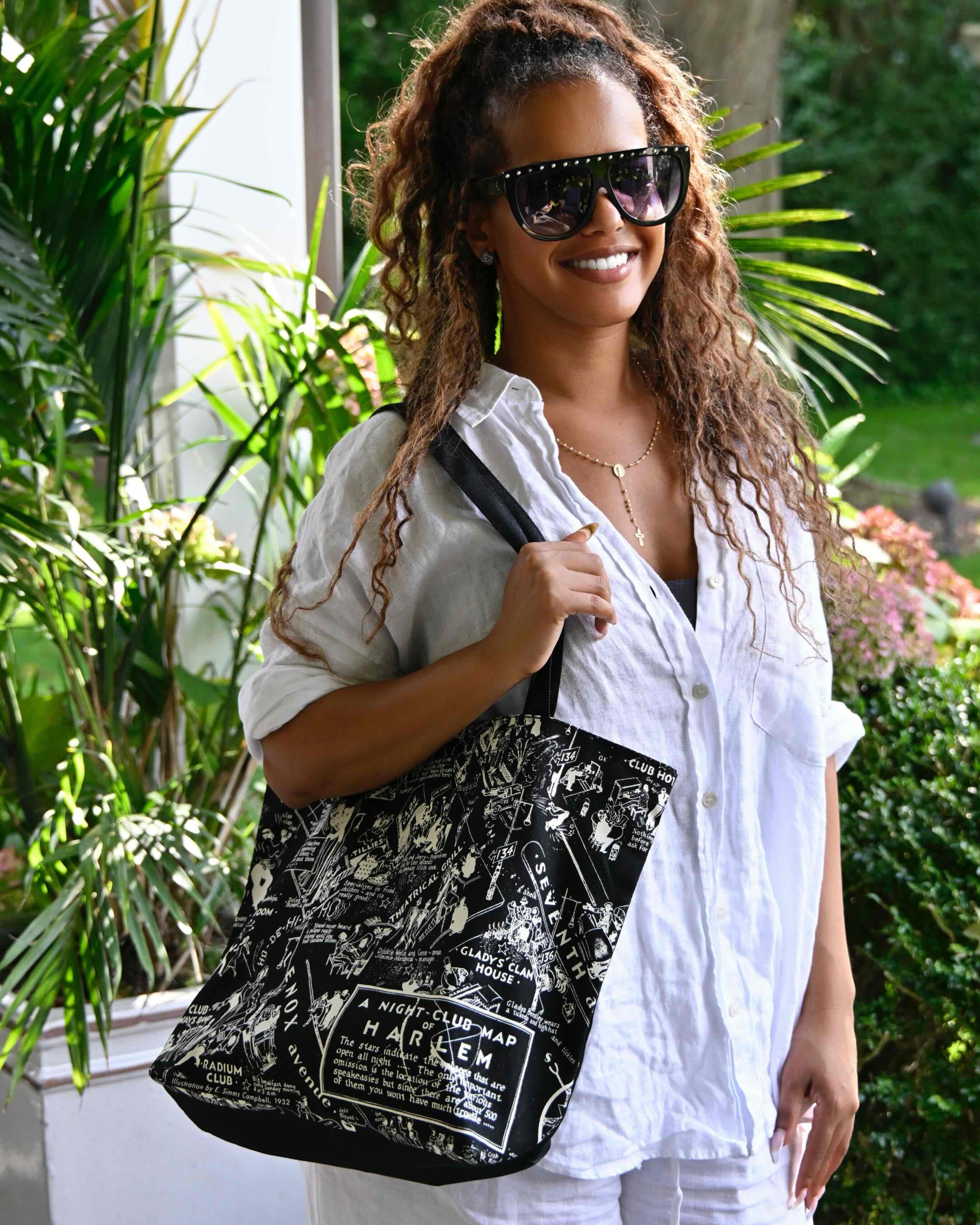 This is an image of a model dressed in white with sunglasses showcasing the black and white Nightclub Map of Harlem tote bag.