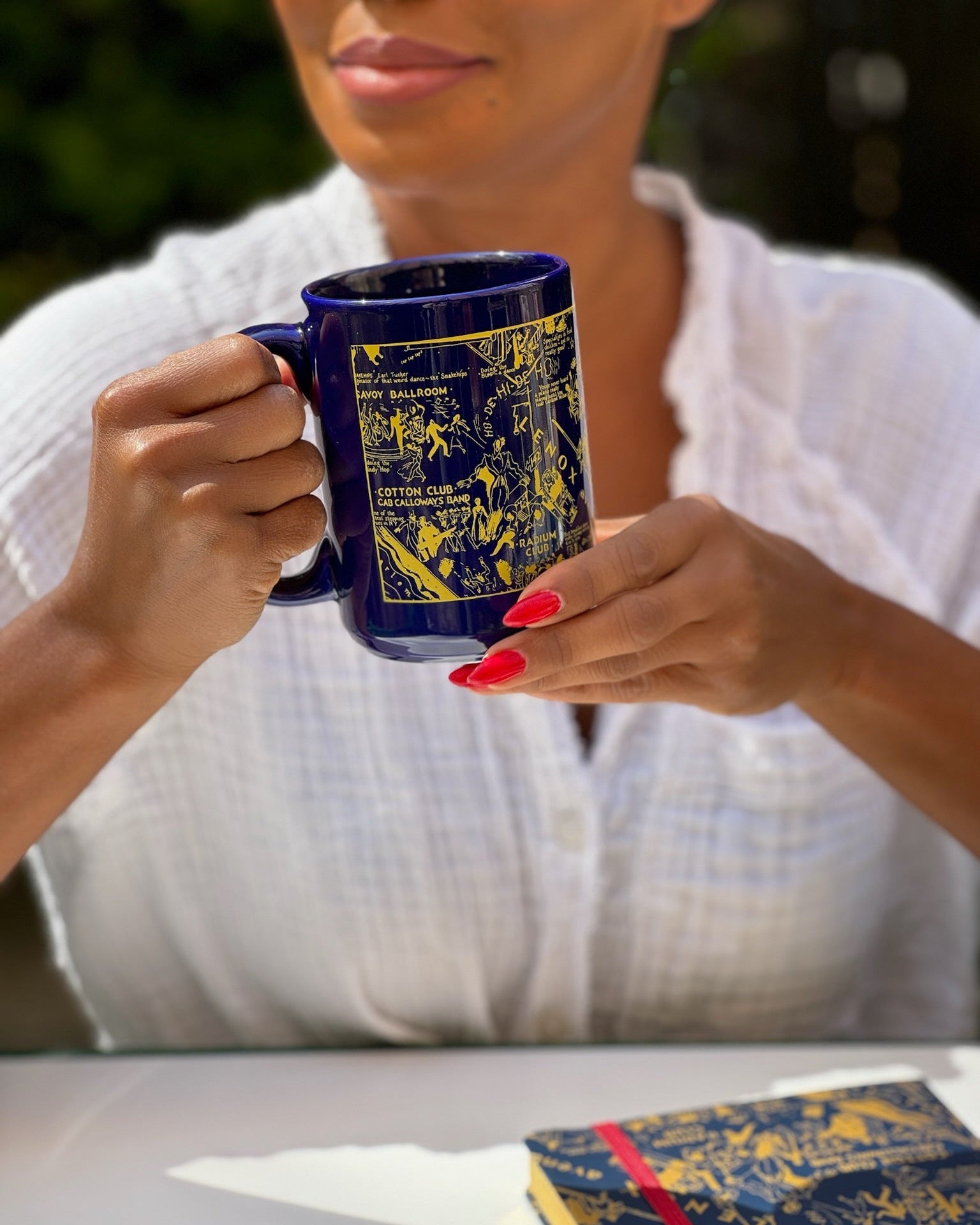 This is a lifestyle image of our blue Nightclub Map of Harlem mug held by a model with red fingernails . She is sitting in front of the blue nightclub map of Harlem journal
