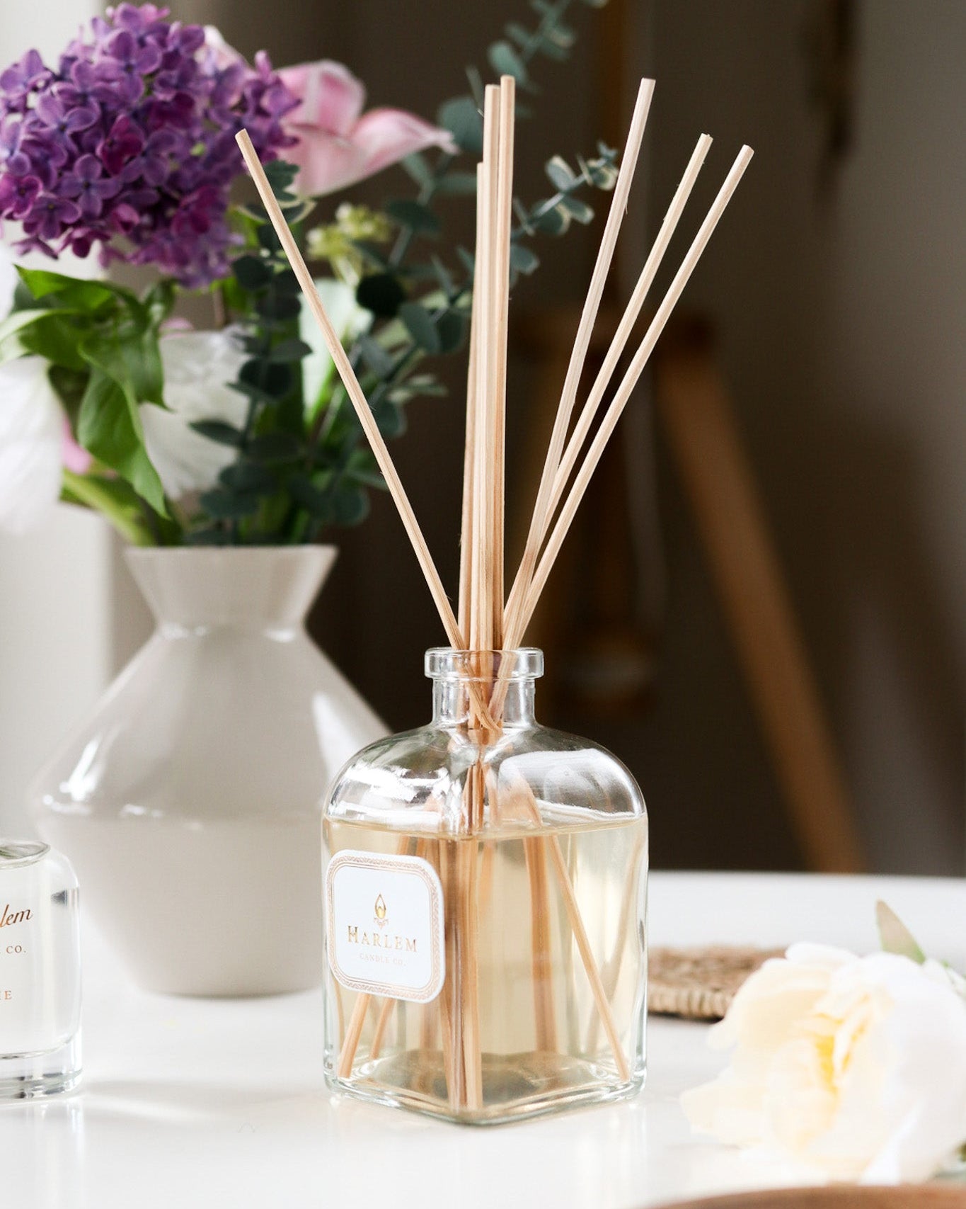 This is a vertical image of the clear glass "Home" Botanical Diffuser bottle with colorful flowers in the background.