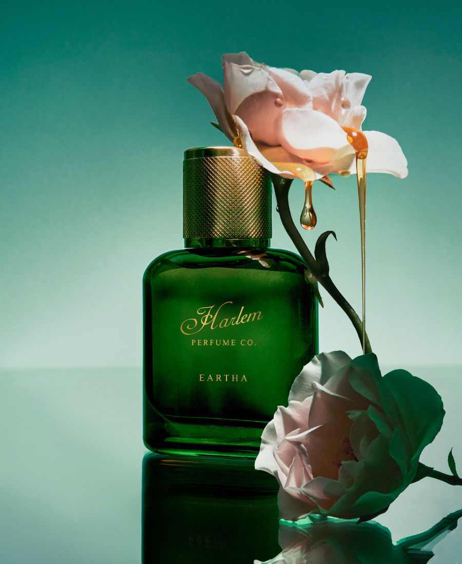 Green perfume bottle with gold cap labeled 'Harlem Perfume Co' next to pink roses on a reflective surface.