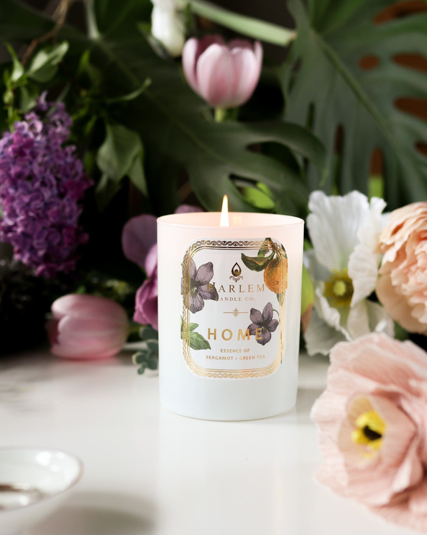 This is a vertical life style image of the "Home" Luxury Candle with flowers in the background.