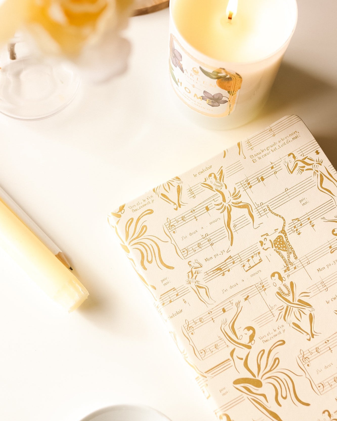 This is a lifestyle image of the cream and gold. Josephine journal featured next to our home candle in a white glass with a floral and white label.