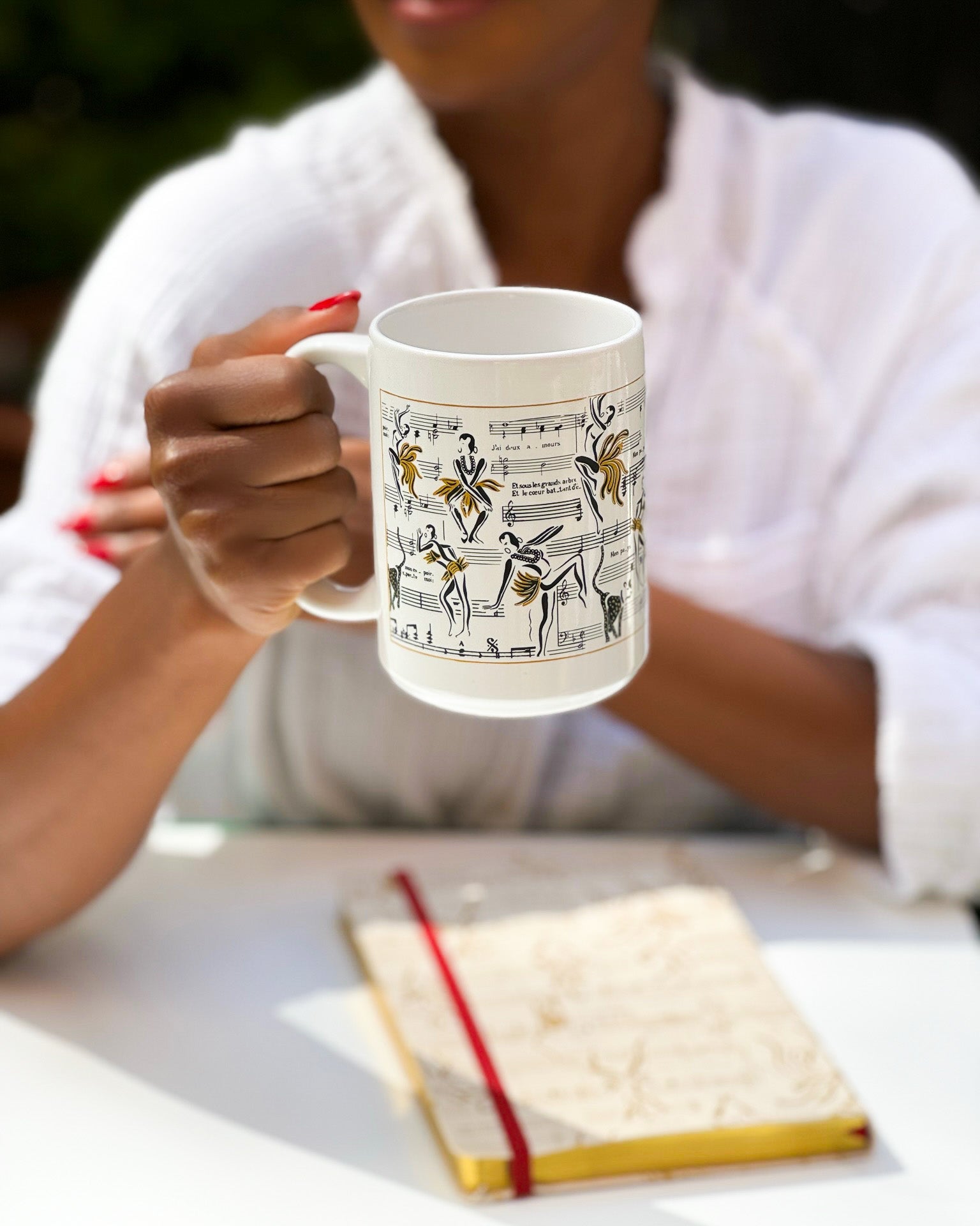 This is an image of the white Josephine mug with illustrations of Josephine Baker dance with her music notes in the background. The Josephine journal is also featured in this image.