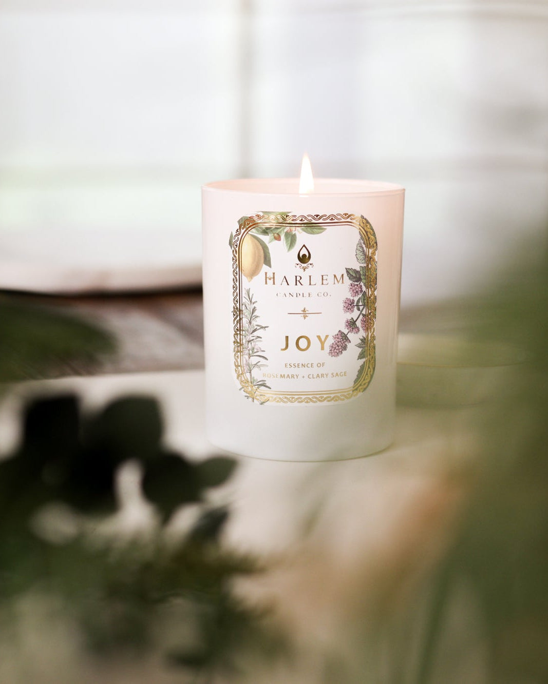 This is an image of the "Joy" Luxury Candle on a wooden table with a white place mat and greenery in the foreground.
