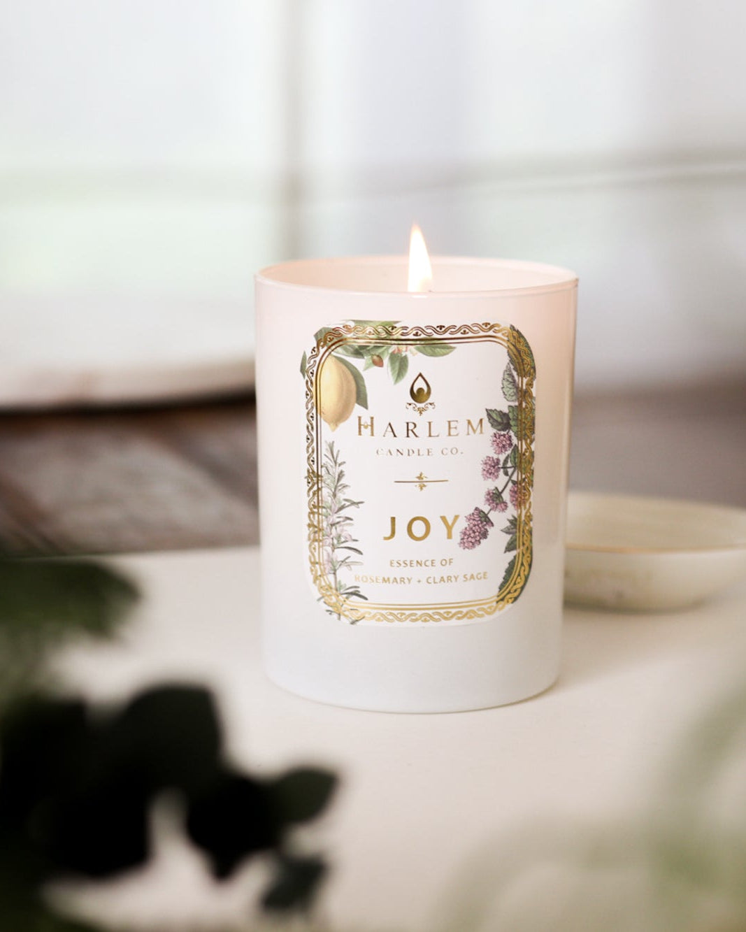 This is an image of the "Joy" Luxury Candle on a wooden table with a white place mat and greenery in the foreground.