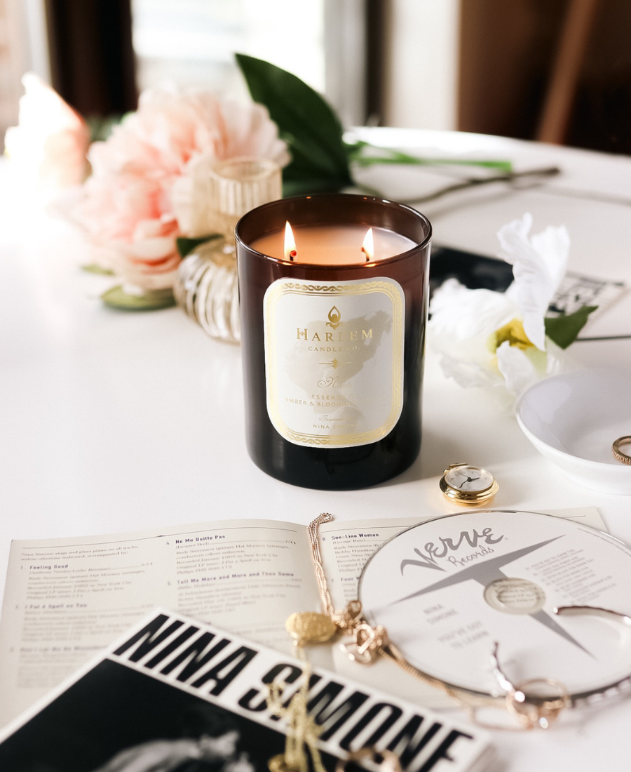 Nina candle on a white table with jewelry, flowers, and a Nina Simon CD and pamphlet.