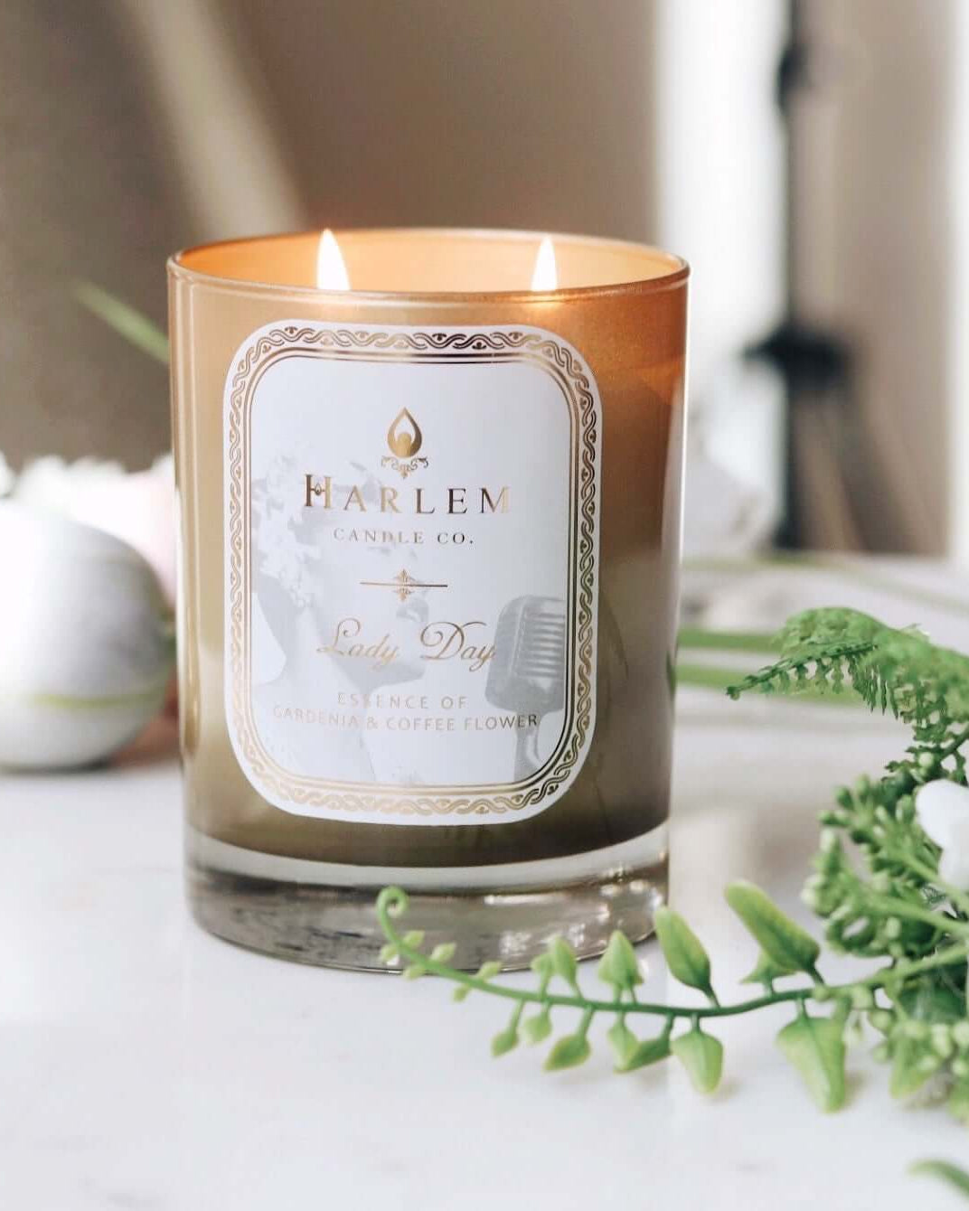 Gold and White Lady Day candle with jasmine, coffee flower, coconut and sandalwood scents.  This candle has 2 wicks.  It is in a lifestyle setting  next to white flowers.