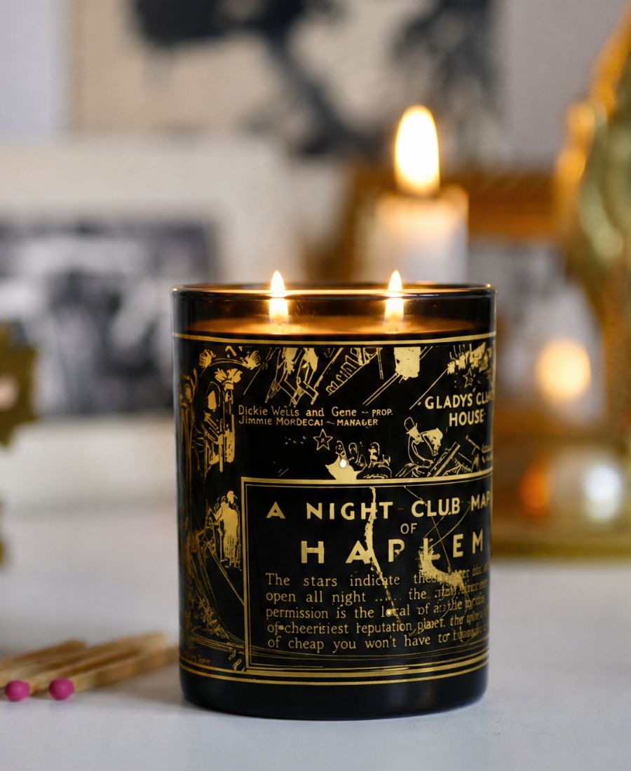 A lit black glass candle jar with gold illustrated text and design, reading 'A Night Club Man of Happin', set on a light surface with a blurred indoor background and warm lights.