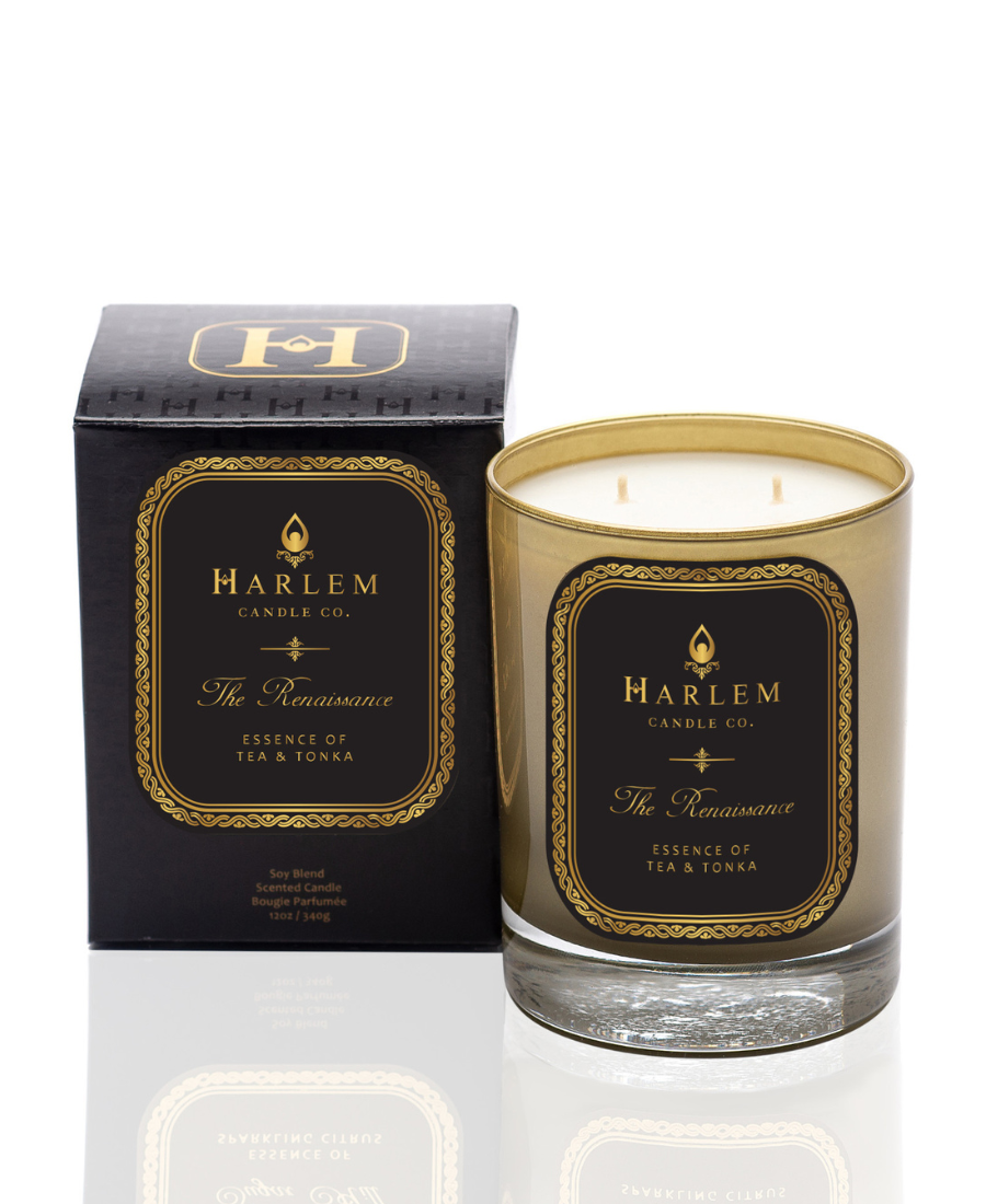 This is an image of The Renaissance Candle pictured in a gold glass with a black and gold label. It is positioned next to its decorative box.