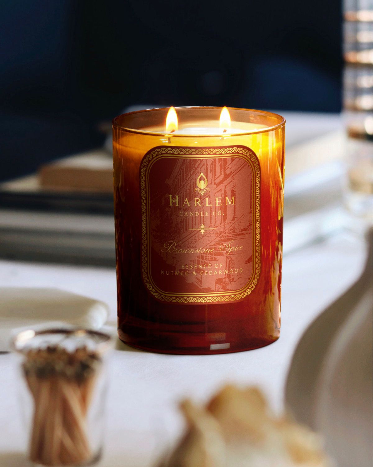 This is a lifestyle image Brownstone Spice 11 oz candle in a burnt orange glass with two flames.