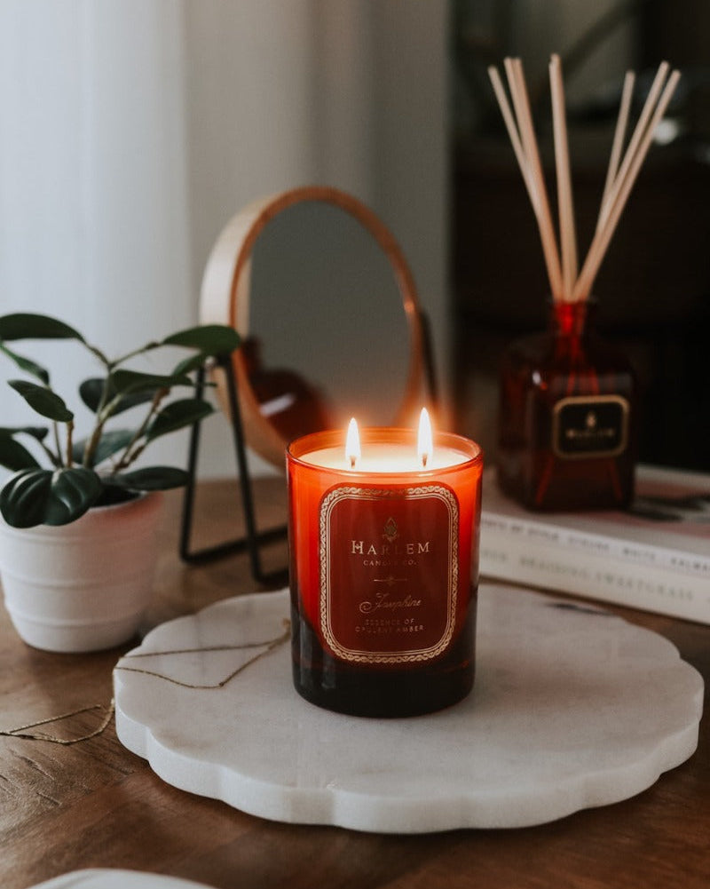 Our 11oz. 2-wick Josephine Candle in a red glass vessel sitting on a wooden table, next to a plant with the Josephine diffuser in the background.