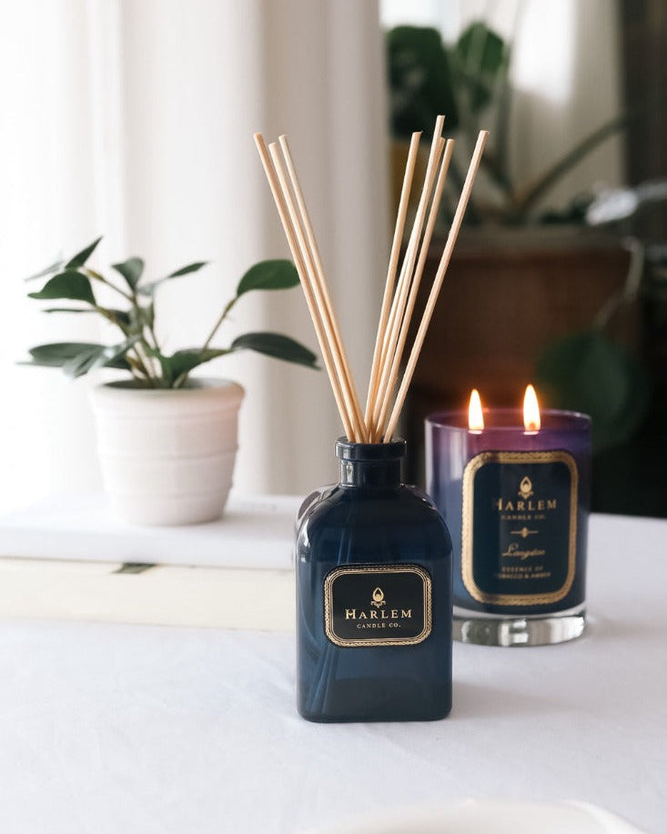 Our 4 fluid oz Langston Reed Diffuser with blue glass and reeds placed in diffuser glass sitting on a white table next to a 11oz Langston Candle.