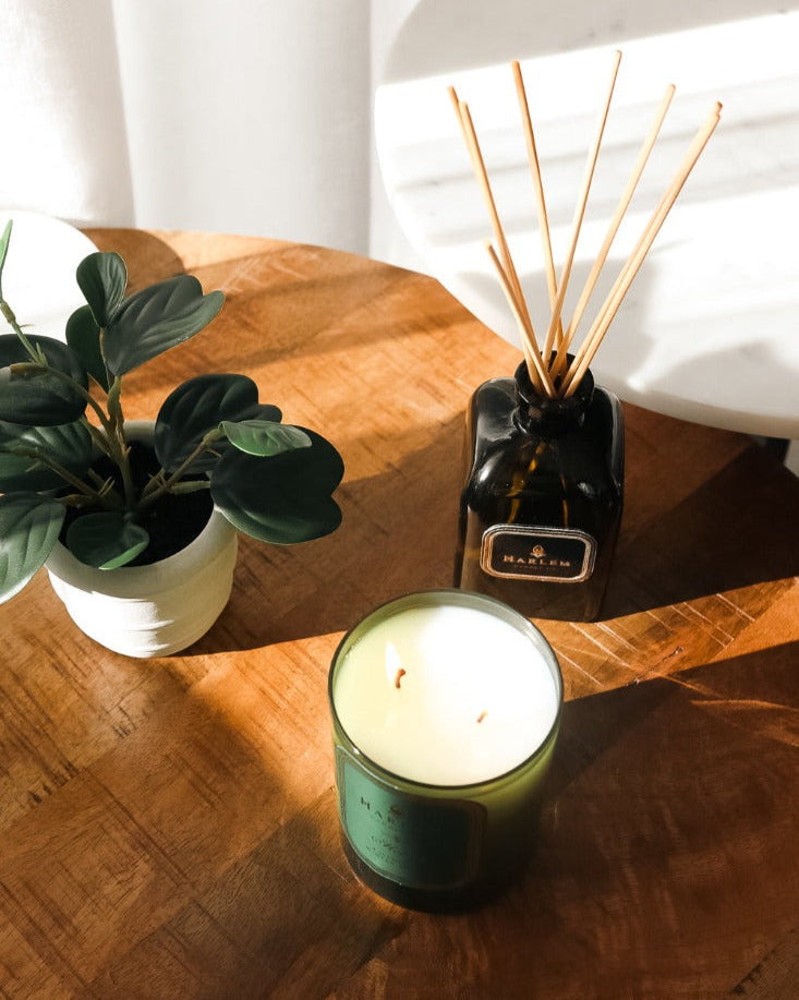 Our 8 fluid oz. Holiday Reed Diffuser with reeds, in a green glass vessel next to a green Holiday 11oz candle. sitting on a brown table..