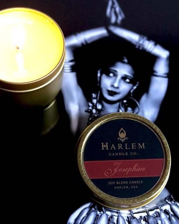 Our Josephine 4 oz travel candle in tin with top next to the soft glowing candle on a black background with a black and white image of Josephine Baker.