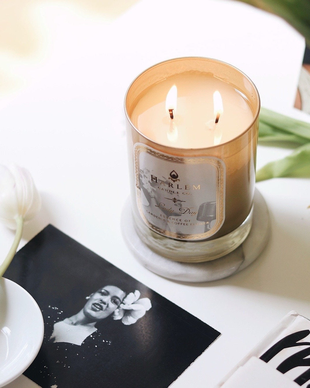 This is a Lifestyle image of the Lady Day candle that is softly glowing on a table with black and white photographs of Billie Holiday and a vase of flowers.