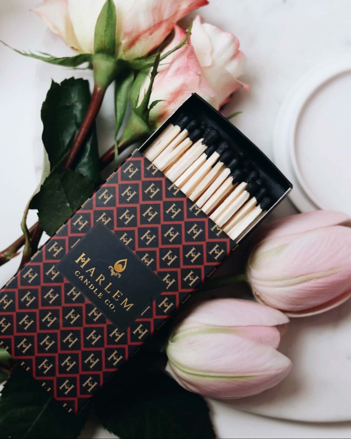 Red, black and gold Match box containing 3 inch matches with black tips.  The box has the Harlem H pattern in an art deco style printed on the box.  The image is on top of flowers.