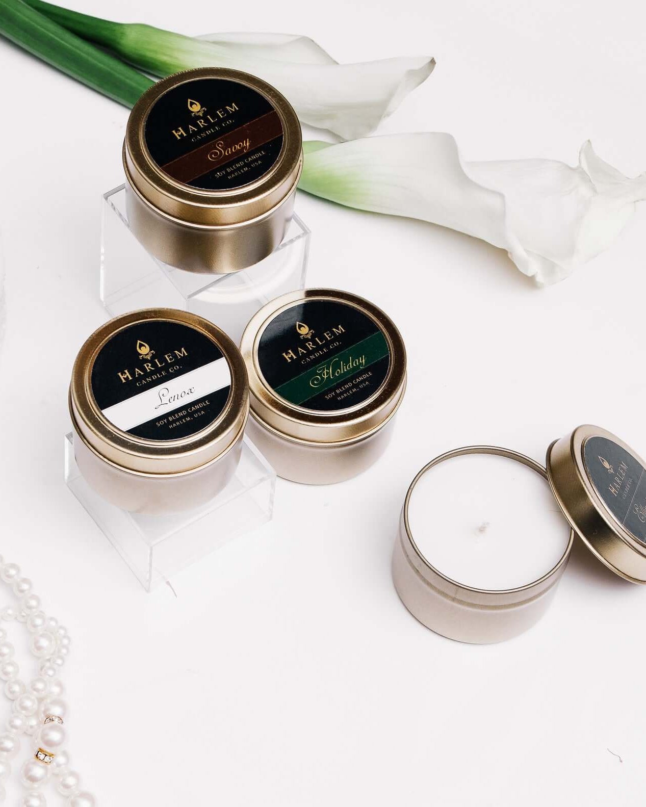 This photo is of our Savoy Luxury Travel Candle 4 oz. Scented with lily of the valley, pear and apple surrounded by a jewelry box with pearls and other travel candles on a white table.