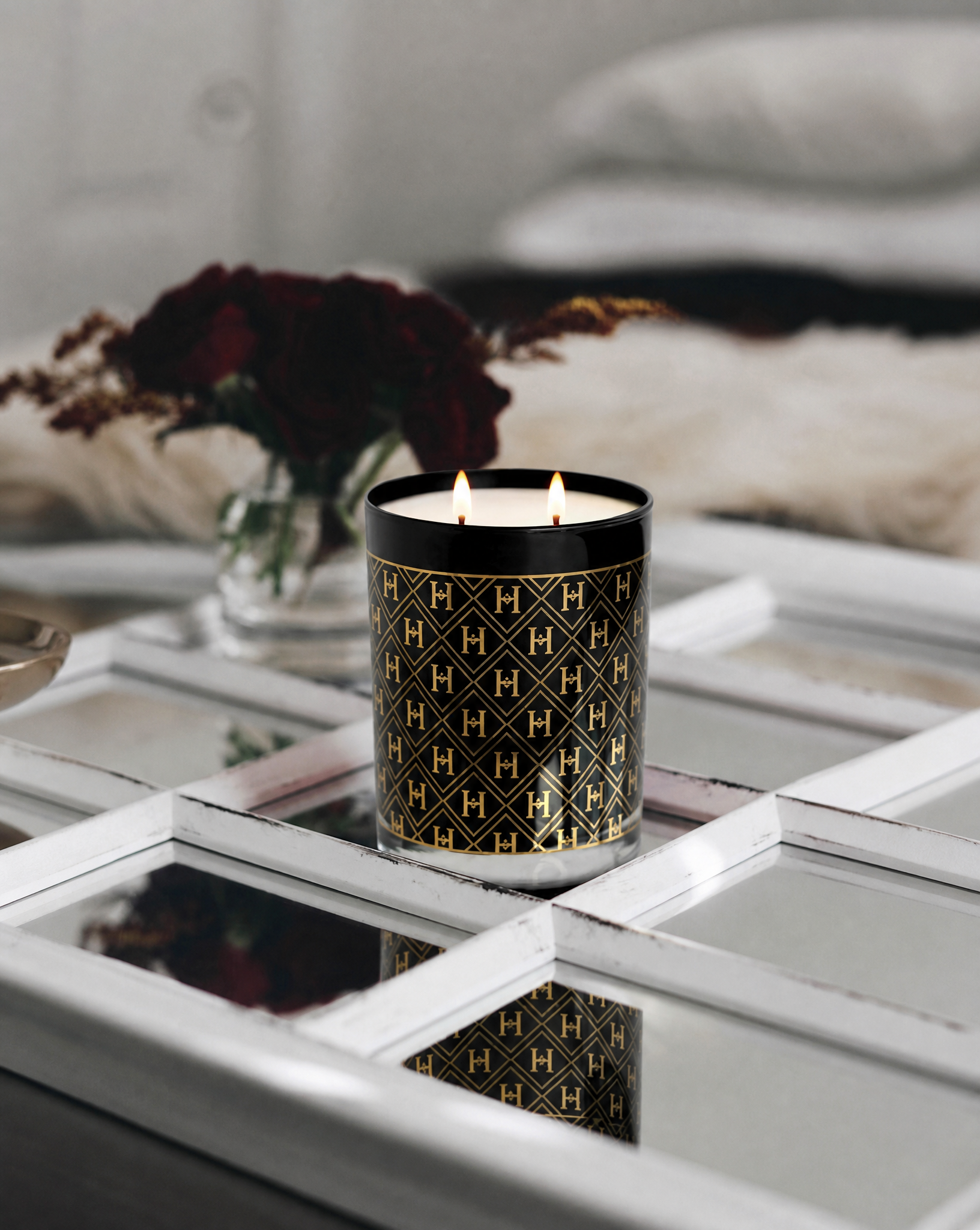 A black double-wick candle with a gold geometric monogram pattern sits on a mirrored tray in a styled interior setting with a blurred vase of dark red flowers in the background.