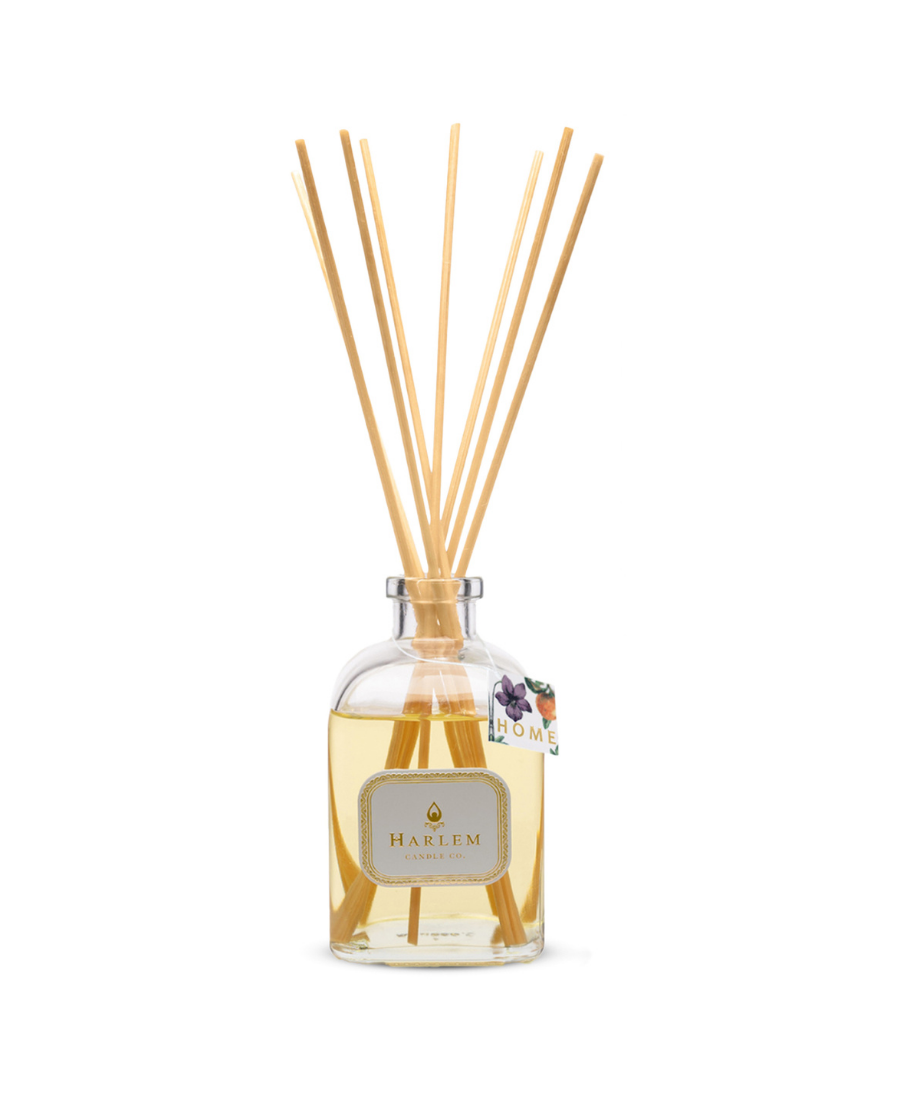 Home reed diffuser bottle with tag and multiple natural wood reed sticks rising above the top, set against a plain white background.