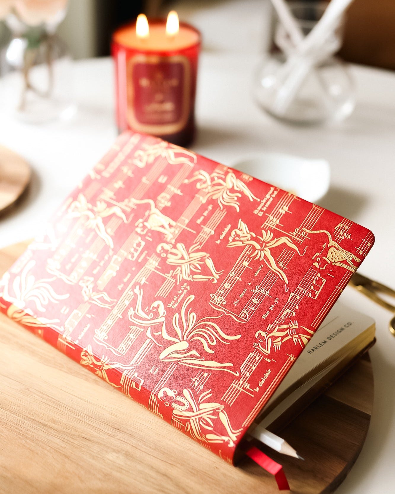 Lifestyle picture of the red Josephine journal on a wood table with the Josephine candle burning in the background. 