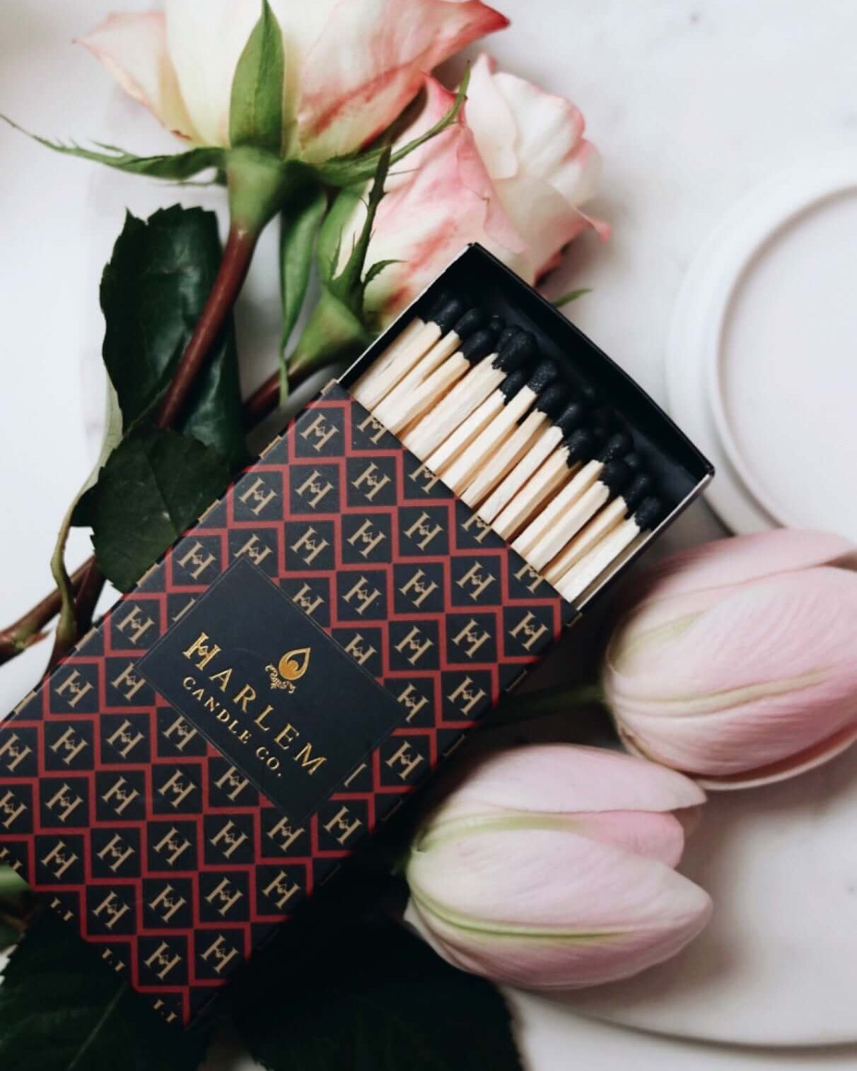 Red, black and gold Match box containing 3 inch matches with black tips.  The box has the Harlem H pattern in an art deco style printed on the box.  The image is on top of flowers.
