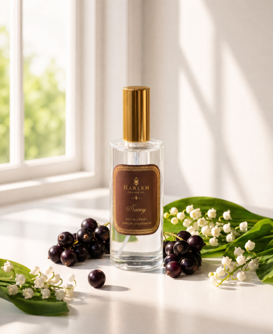 A clear glass perfume bottle with a gold cap and a brown label reading 'HARLEM styled on a windowsill with white flowers, green leaves, and dark berries in soft natural light.