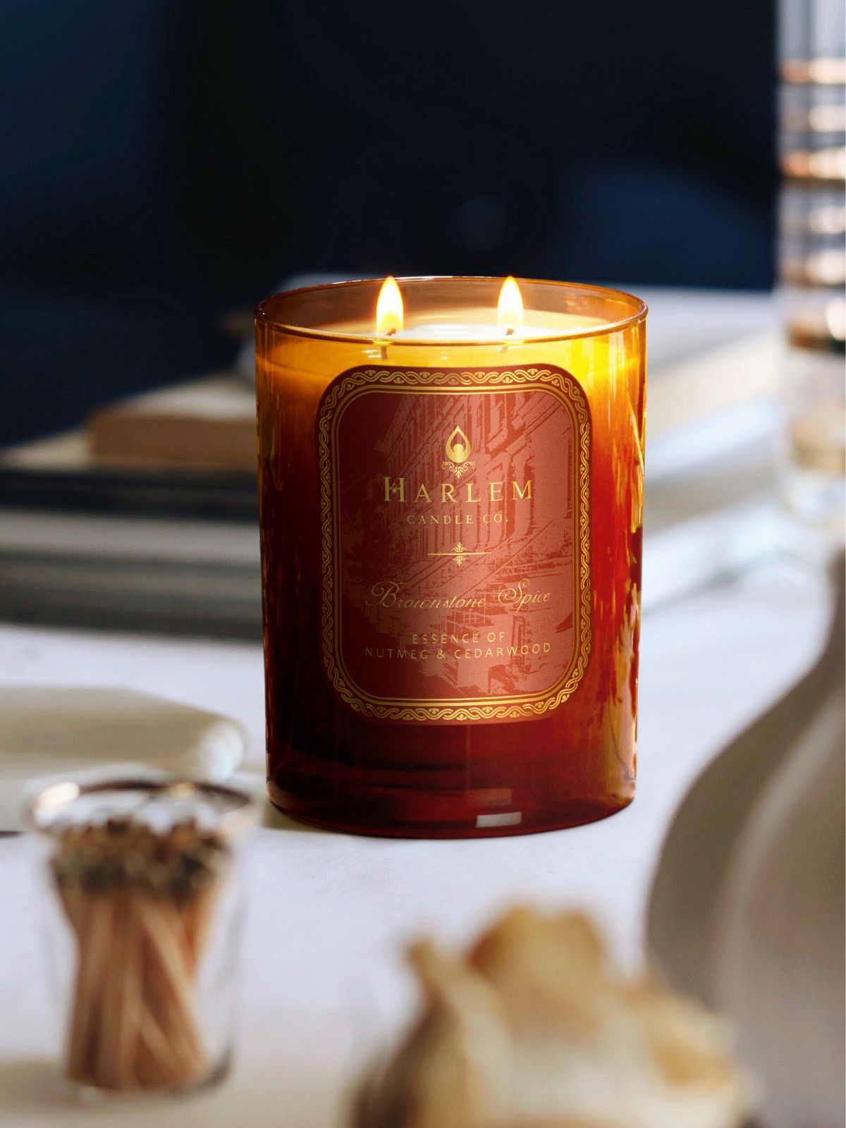 This is a lifestyle image Brownstone Spice 11 oz candle in a burnt orange glass with two flames.