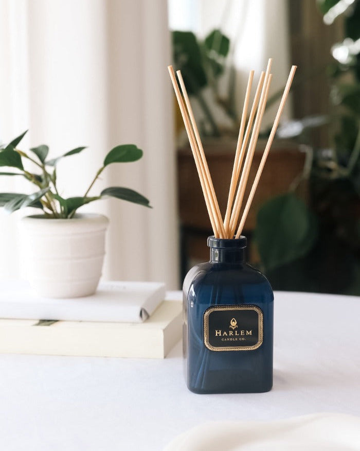 Our 4 fluid oz Langston Reed Diffuser with blue glass and reeds placed in diffuser glass sitting on a white table next to a plant.