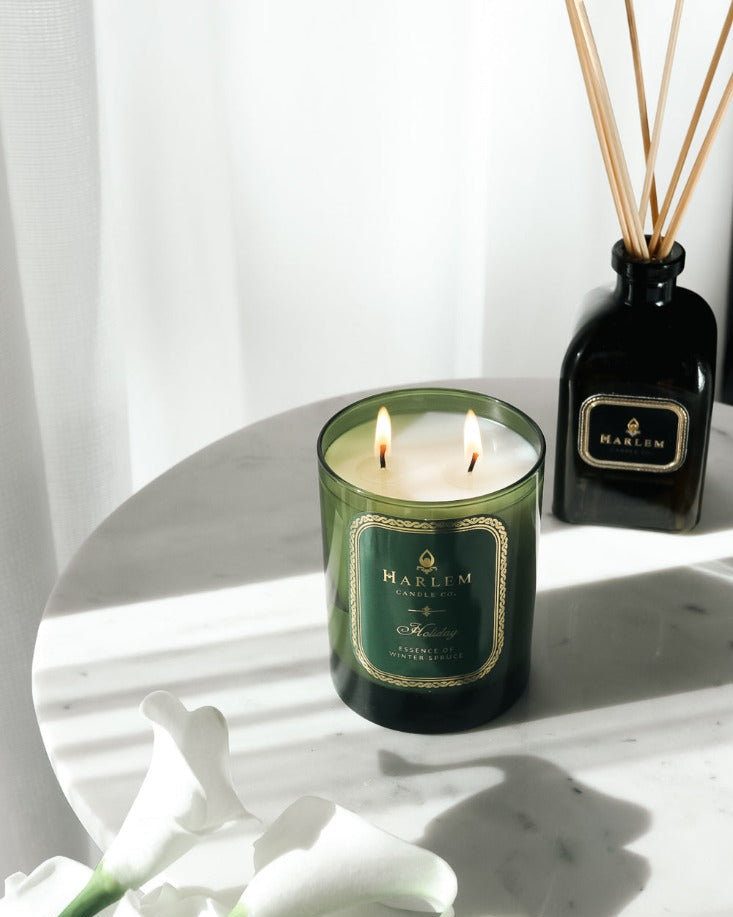 Our 8 fluid oz. Holiday Reed Diffuser with reeds, in a green glass vessel next to a green Holiday 11oz candle. sitting on a white table