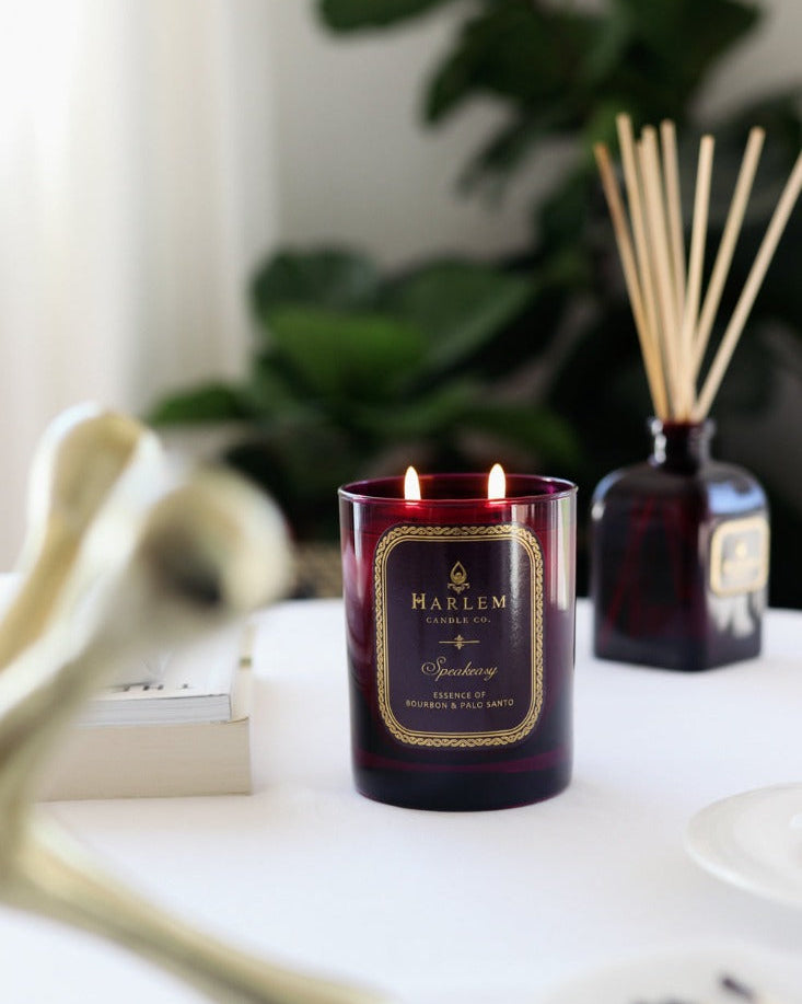 Our 8 fluid oz. Speakeasy Reed Diffuser with reeds,and 11oz Speakeasy Candlein a purple glass vessel sitting next to its decorative box on a white table.