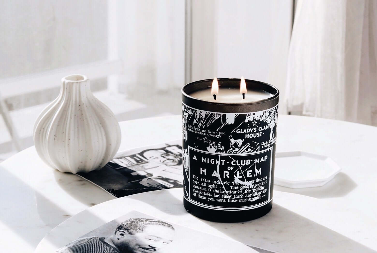 Our black and white Langston Nightclub Map 12 oz, 2 wick candle lit sitting on a white side table with black and white photographs and a beautiful white vase next to the candle.
