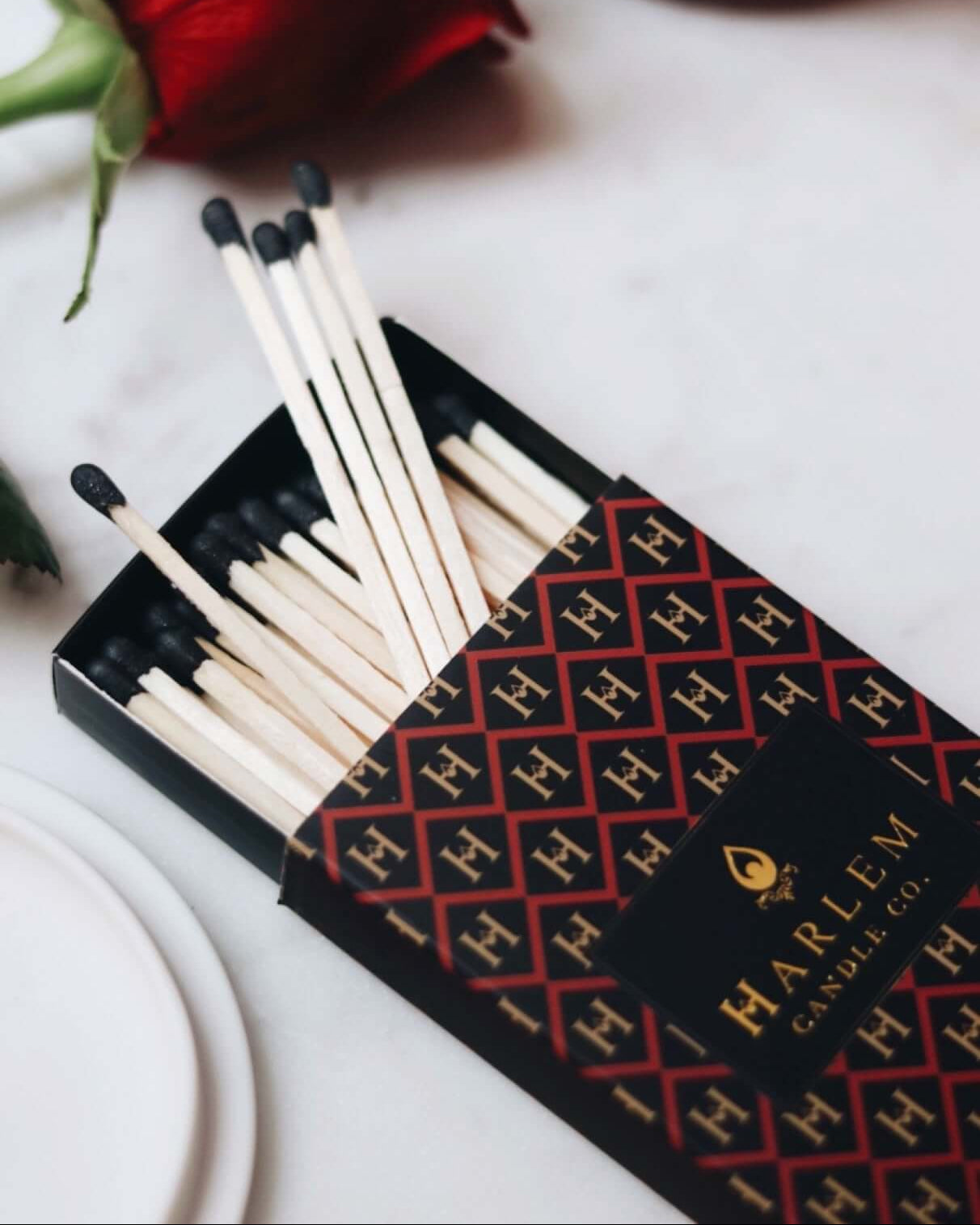 Red, black and gold Match box containing 3 inch matches with black tips.  The box has the Harlem H pattern in an art deco style printed on the box.  The image is on next to flowers.