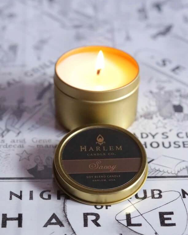 A lifestyle image of our Our stunning Savoy travel candle in a  gold metal tin. The candle has a soft flame and sits atop our black and white Harlem Map.