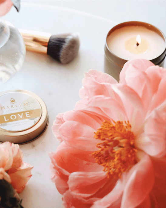 Our gorgeous Love Candle in a metal tin which is softly lit and burning on a table with a vase and flowers.